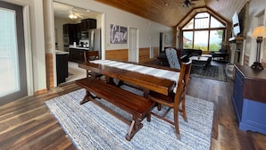 Een ruime eethoek met een houten tafel en banken, een keuken op de achtergrond, en een groot raam met uitzicht op de omgeving.