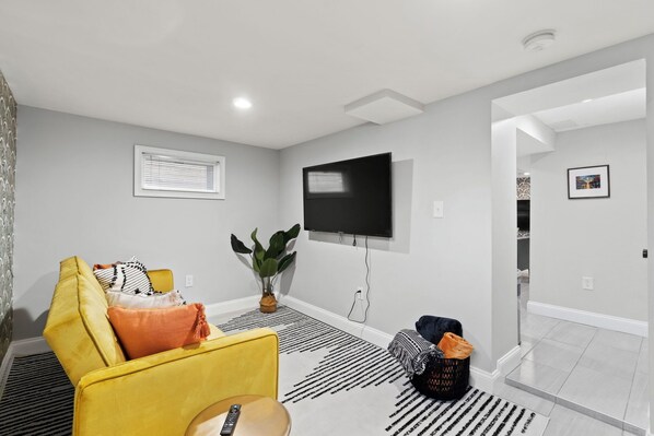 A modern living room with a yellow sofa, a flat-screen TV mounted on the wall, a potted plant, and a striped rug.