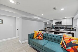 A modern living room with a teal sofa, colorful cushions, and a kitchen in the background.