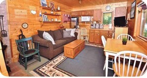 Interior de una cabaña de madera con sofá, sillas, una mesa de comedor, una zona de cocina y una pequeña alfombra.