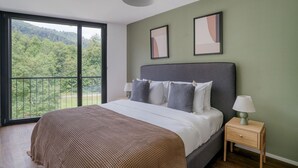 A bedroom with a large bed, a nightstand, a lamp, and a view of a forest through a balcony. - Chic Wollishofen 1BR w/ Storage & W/D, nr Local Market, by Blueground (Zurich)