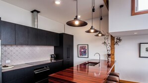 A modern kitchen with dark cabinetry, a wooden island with stools, pendant lighting, and artwork on the walls. - The Thomas Gregg Apartments (Dunedin)