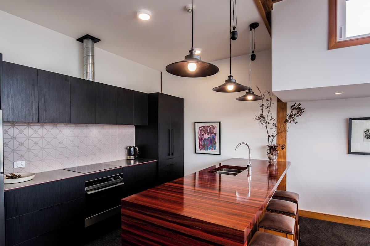 A modern kitchen with dark cabinetry, a wooden island with stools, pendant lighting, and artwork on the walls.