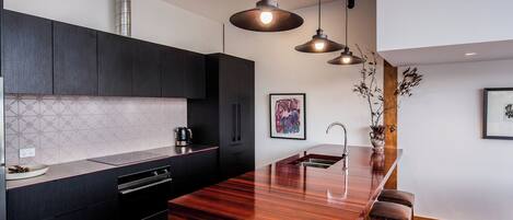 A modern kitchen with dark cabinetry, a wooden island with stools, pendant lighting, and artwork on the walls.