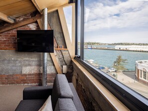 A modern room with a large window overlooking a waterfront, a fireplace, and a flat-screen TV mounted on the wall. - The Thomas Gregg Apartments (Dunedin)