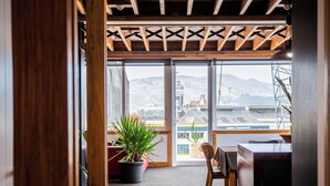 A room with a wooden ceiling, a large window, a potted plant, a chair, and a table. - The Thomas Gregg Apartments (Dunedin)