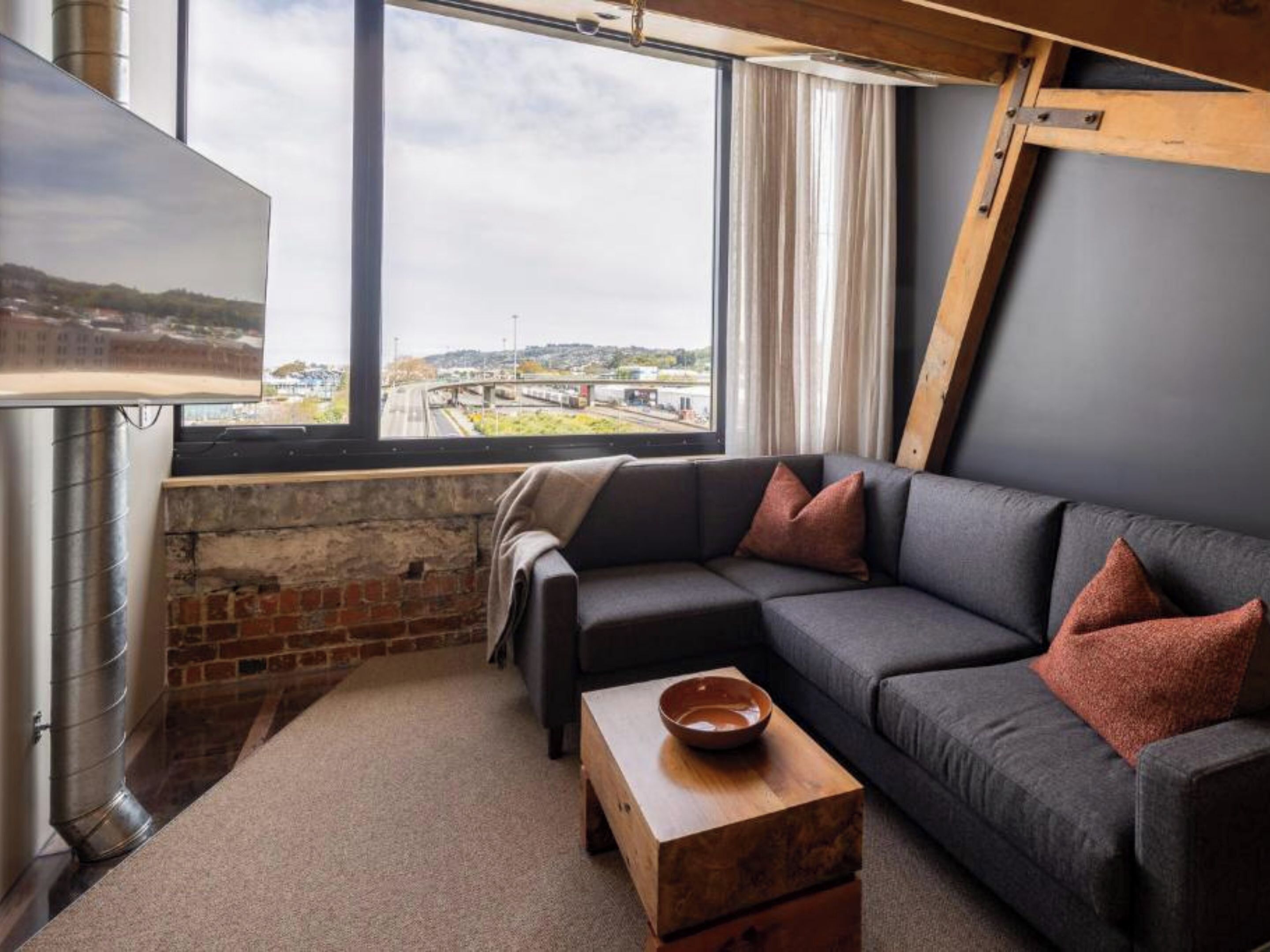 A modern living room with a gray sectional sofa, a wooden coffee table, and a large window offering a view of a bridge and landscape.