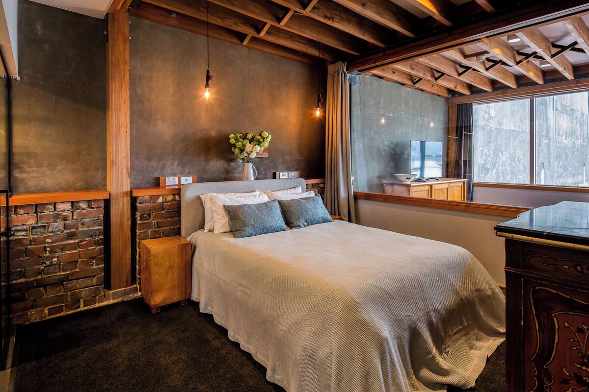 A bedroom with a brick wall, wooden beams, a bed with pillows, a nightstand, a vase with flowers, and a window with a view.