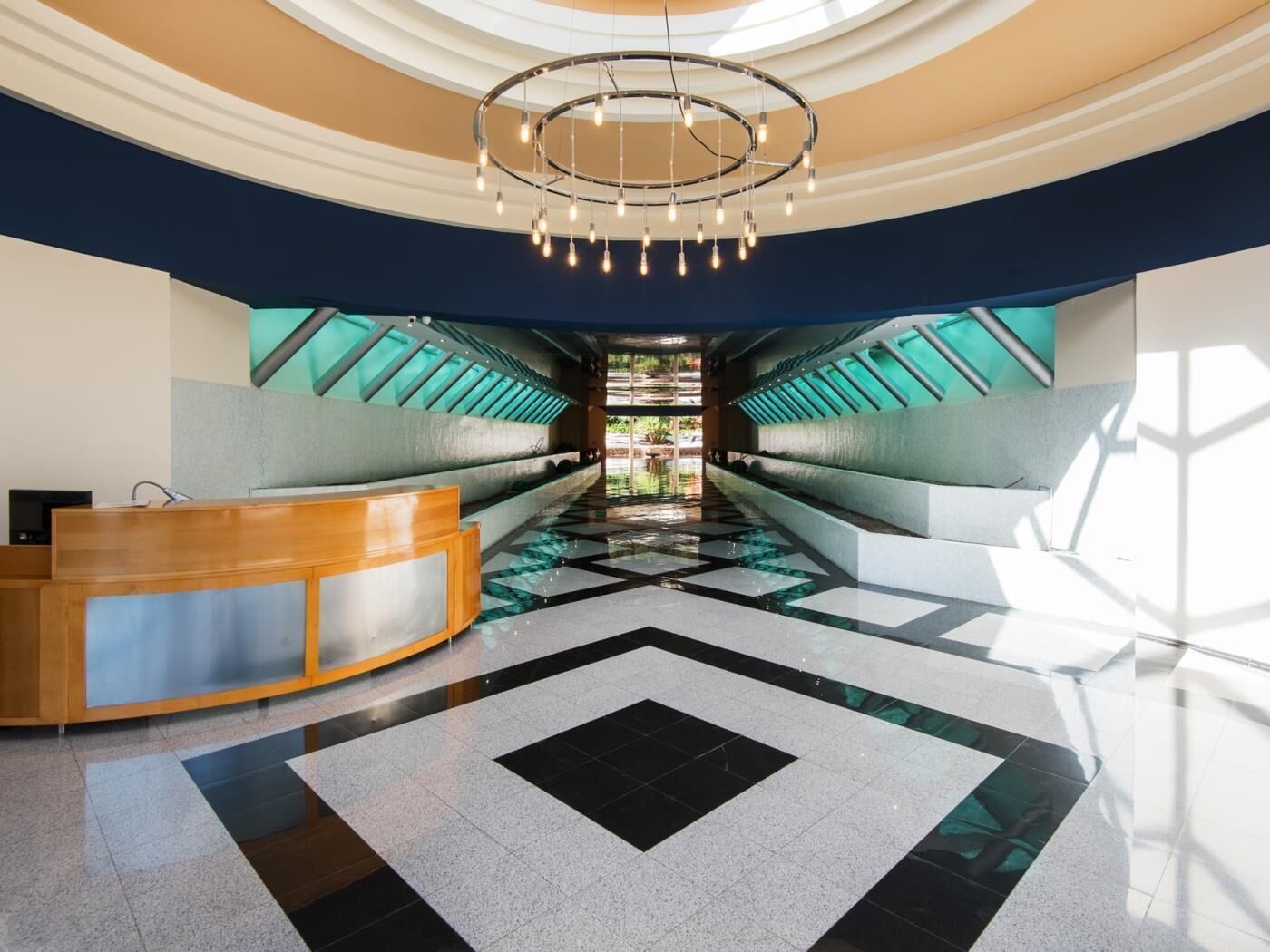 A modern hotel lobby with a central reception desk, a geometric patterned floor, and a large circular chandelier.