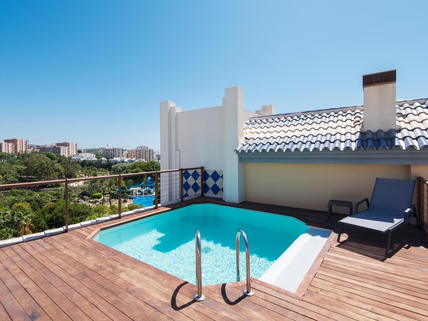 A rooftop pool with a wooden deck, a view of the city, and a white building with blue tiles.
