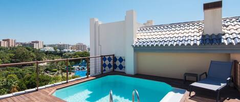 A rooftop pool with a wooden deck, a view of the city, and a white building with blue tiles.
