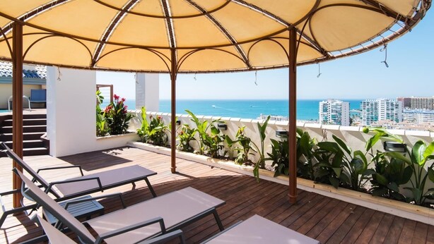 Un espacio al aire libre con mesas de madera y sillones, rodeado de plantas, con vistas a la ciudad y al mar.