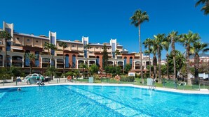 Una piscina con agua cristalina y azul, rodeada por un hotel con varios balcones y palmeras.