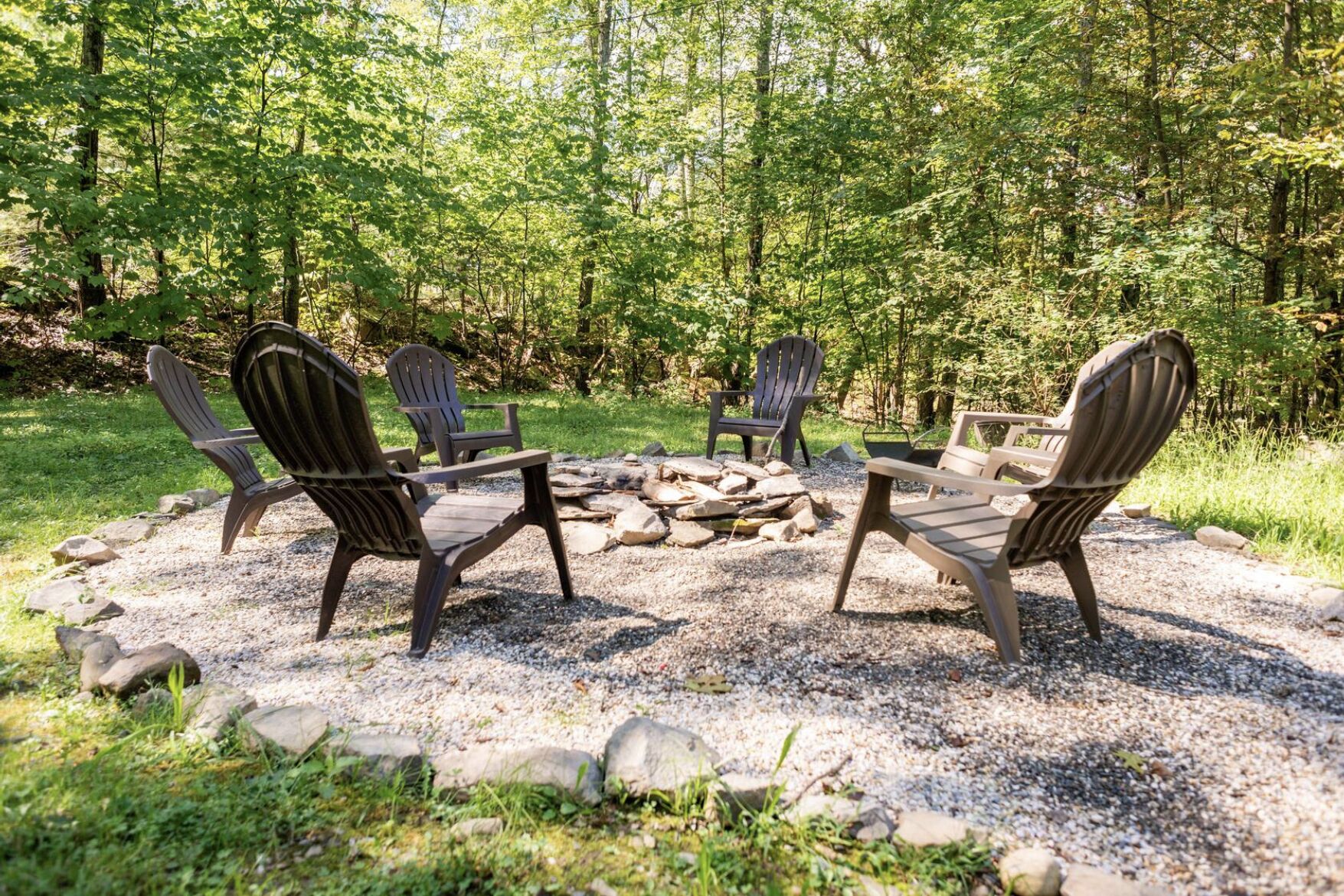 A circular seating area with four Adirondack chairs around a fire pit, set in a forested area with gravel ground cover.