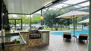 Un área de cocina al aire libre con encimera de piedra, una parrilla y vista a una piscina.