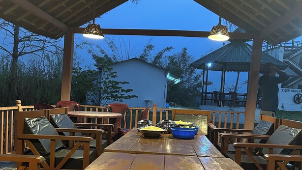 An outdoor dining area with wooden tables and chairs, a covered roof, and a view of a building in the background. - Tiger Wildlife Resort (Jagatpur)
