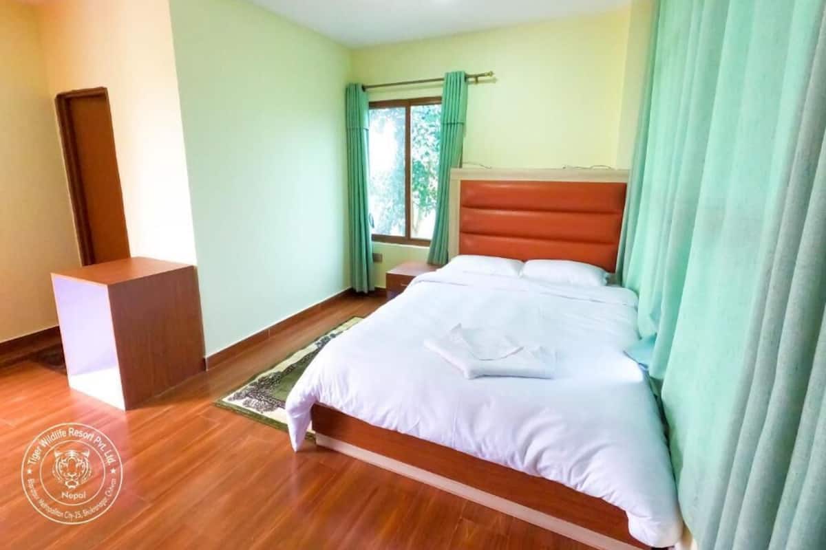 A hotel room with a wooden floor, a bed with white bedding, a wooden headboard, and a window with green curtains.