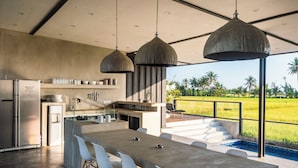 A modern kitchen with a large island, pendant lights, and a view of a rice field. - Joshua District (Tabanan)