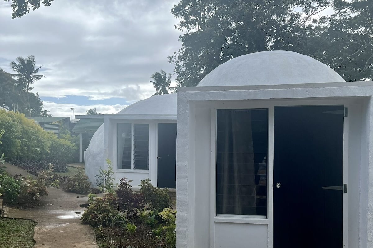 A row of white dome-shaped buildings with large windows and a dark door, surrounded by greenery and a concrete path.