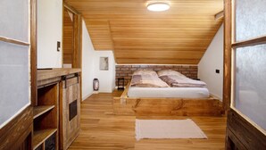 Une chambre à coucher aménagée dans un espace de type « loft », avec un plafond en pente, des poutres apparentes, un mur en briques et un lit agrémenté de linge de lit à motifs.