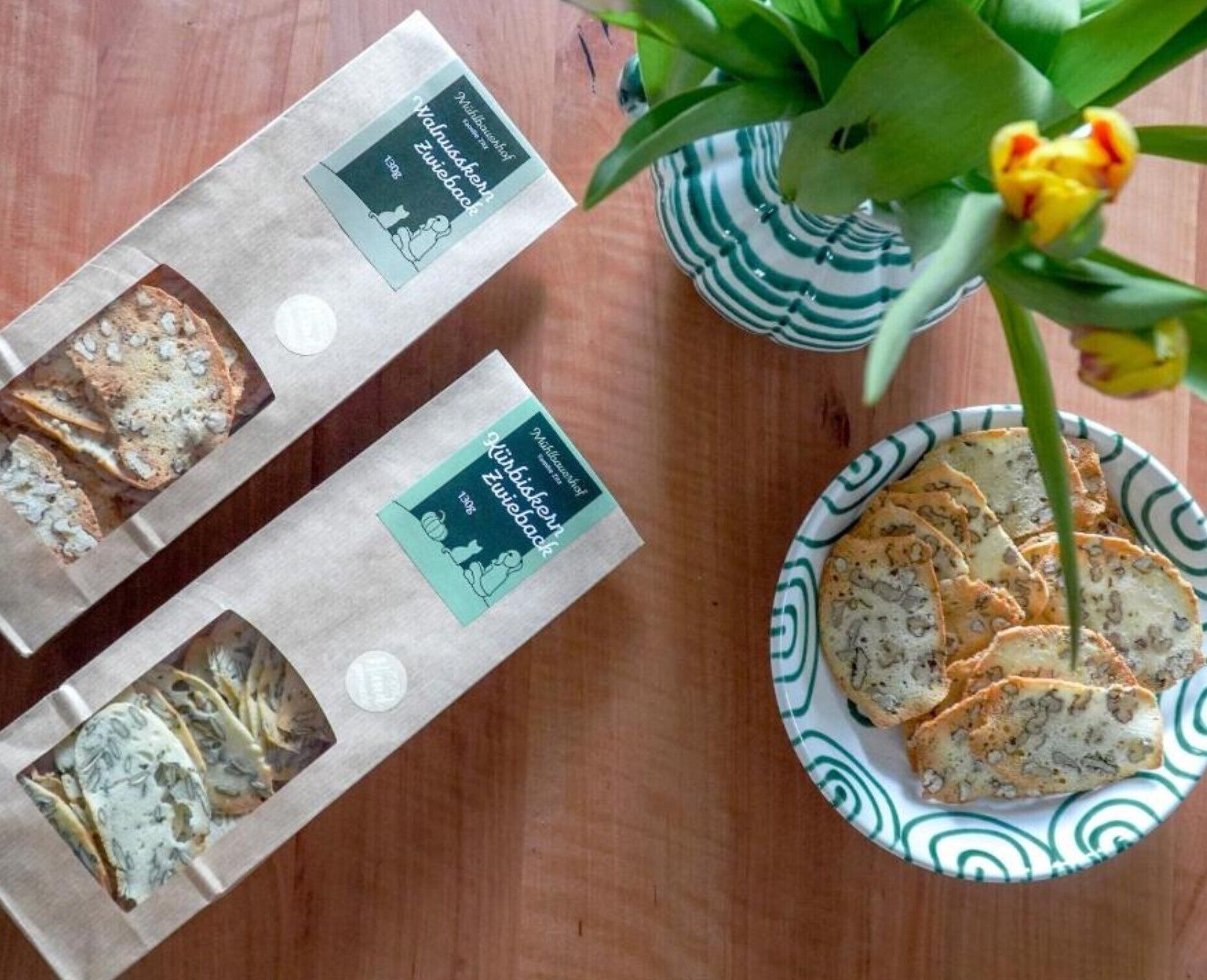 Zwei verpackte Lebensmittel, ein Teller mit Brot und eine Vase mit einer Blume auf einem Holztisch.