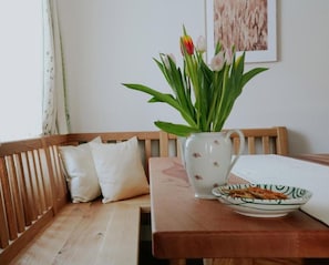 Een houten bank met kussens, een tafel met eten erop en een vaas met bloemen.