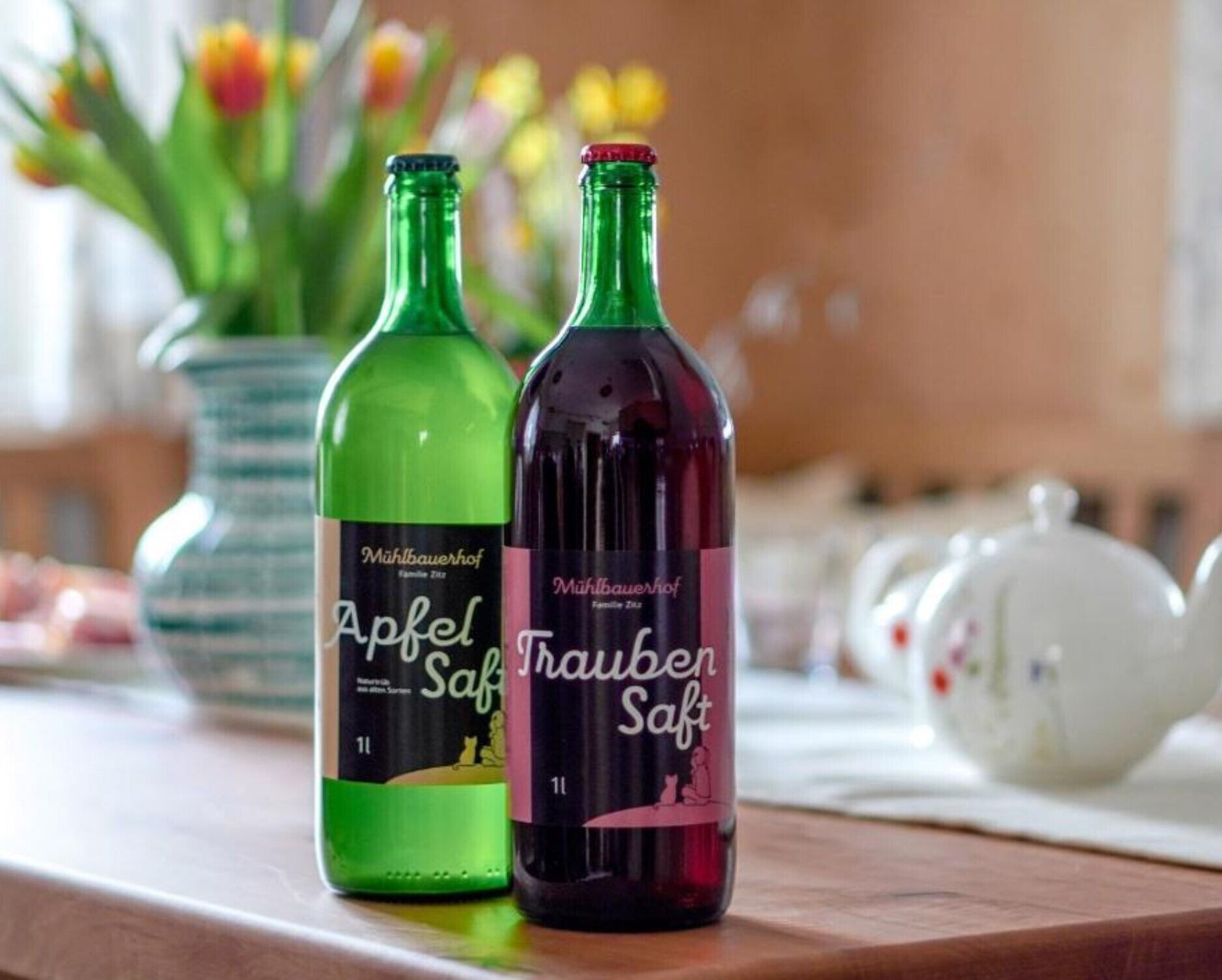 Two bottles of Mühlbauerhof Apfel Saft and Trauben Saft on a wooden surface with a vase of flowers in the background.
