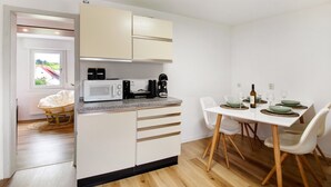 Une cuisine compacte avec des meubles blancs, un four à micro-ondes et une machine à café. On y trouve une table à manger avec des chaises, ainsi qu’un fauteuil en osier dans un coin.