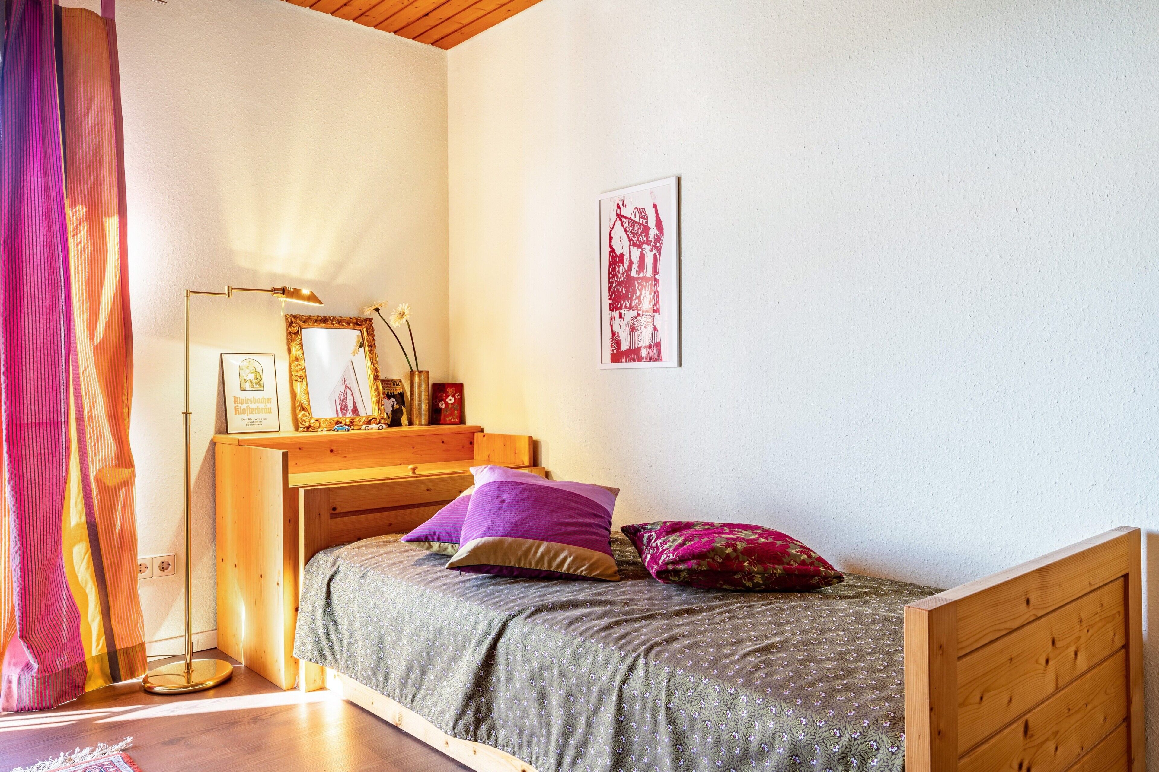 A bedroom with a wooden bed, a bedside table with a lamp, a framed picture, and a window with curtains.