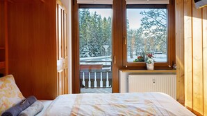 A bedroom with a large window, wooden walls, a bed with floral bedding, and a view of a snowy landscape. - Apartment 'Oskar' with Garden View, 2 Balconies and Wi-Fi (Titisee-Neustadt)