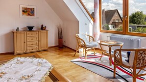 A room with a wooden floor, a dining table with chairs, a sofa, a cabinet, and a window with a view of houses. - Holiday Apartment 'Eule' with Wi-Fi (Donaueschingen)