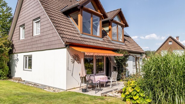 A two-story house with a sloped roof, a balcony with a table and chairs, and a garden area with plants and a hedge. - Holiday Apartment 'Eule' with Wi-Fi (Donaueschingen)