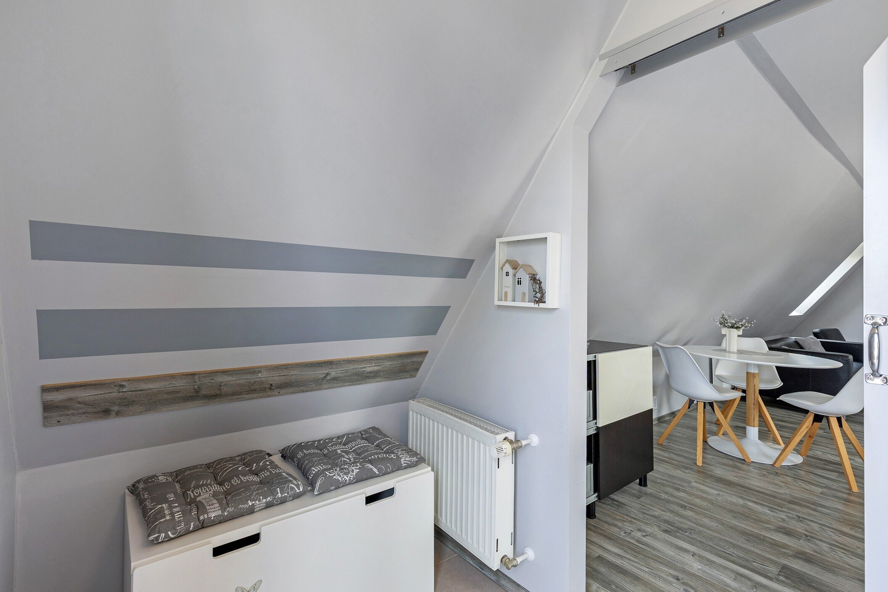 Un dormitorio tipo loft con techo inclinado, un radiador blanco, un banco de madera con cojines, una pequeña mesa blanca con sillas y un cuadro enmarcado en la pared.