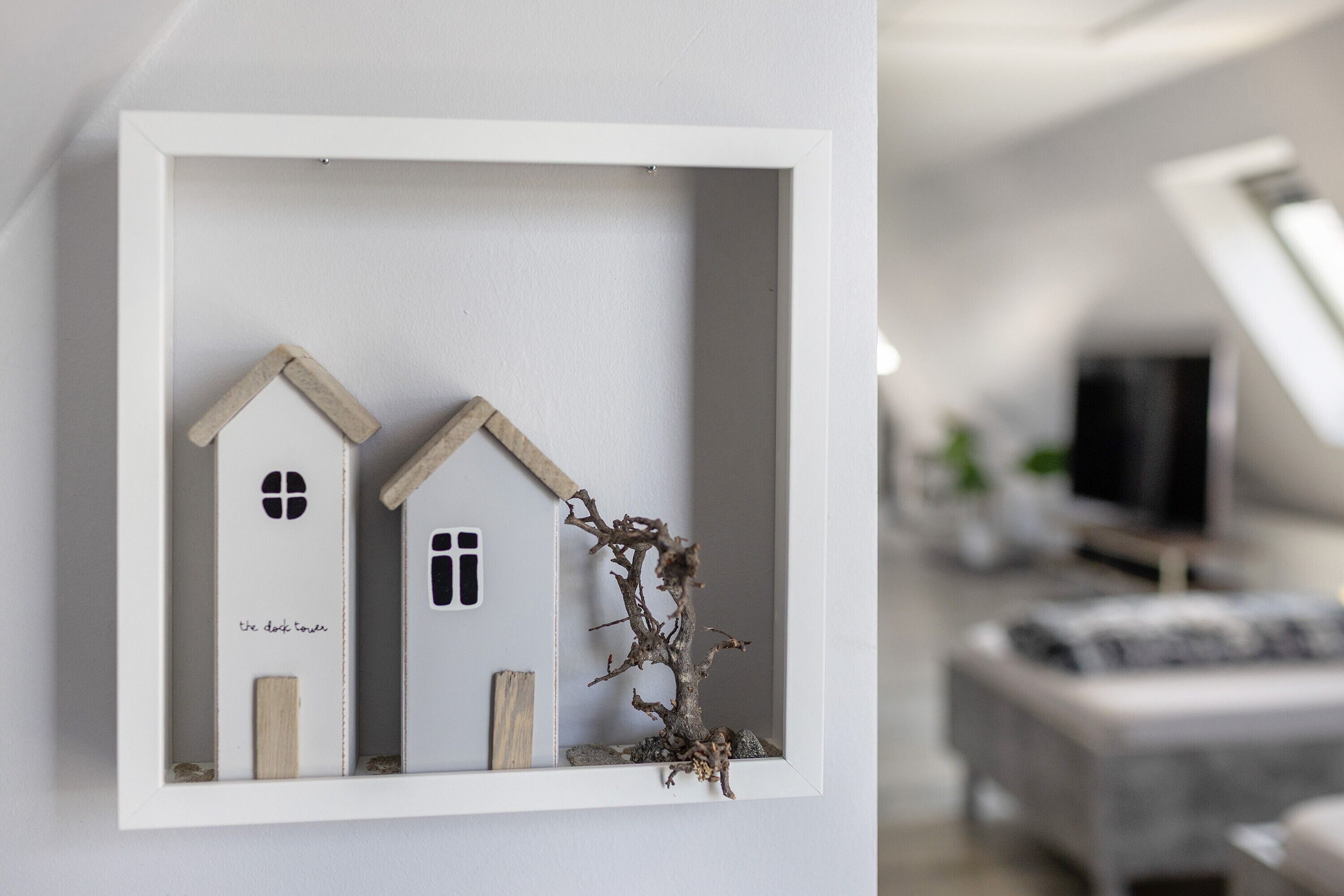 Een miniatuurhuismodel in een lijst met een tak, geplaatst tegen een witte muur in een kamer met een grijze bank en een televisie op de achtergrond.