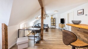 A modern loft-style room with exposed wooden beams, a dining area with a bench, a bar counter, and a seating area with a sofa and a coffee table.