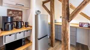 A kitchen with a coffee maker, kettle, and mug on the counter, a refrigerator, wooden beams, and grey cabinets.