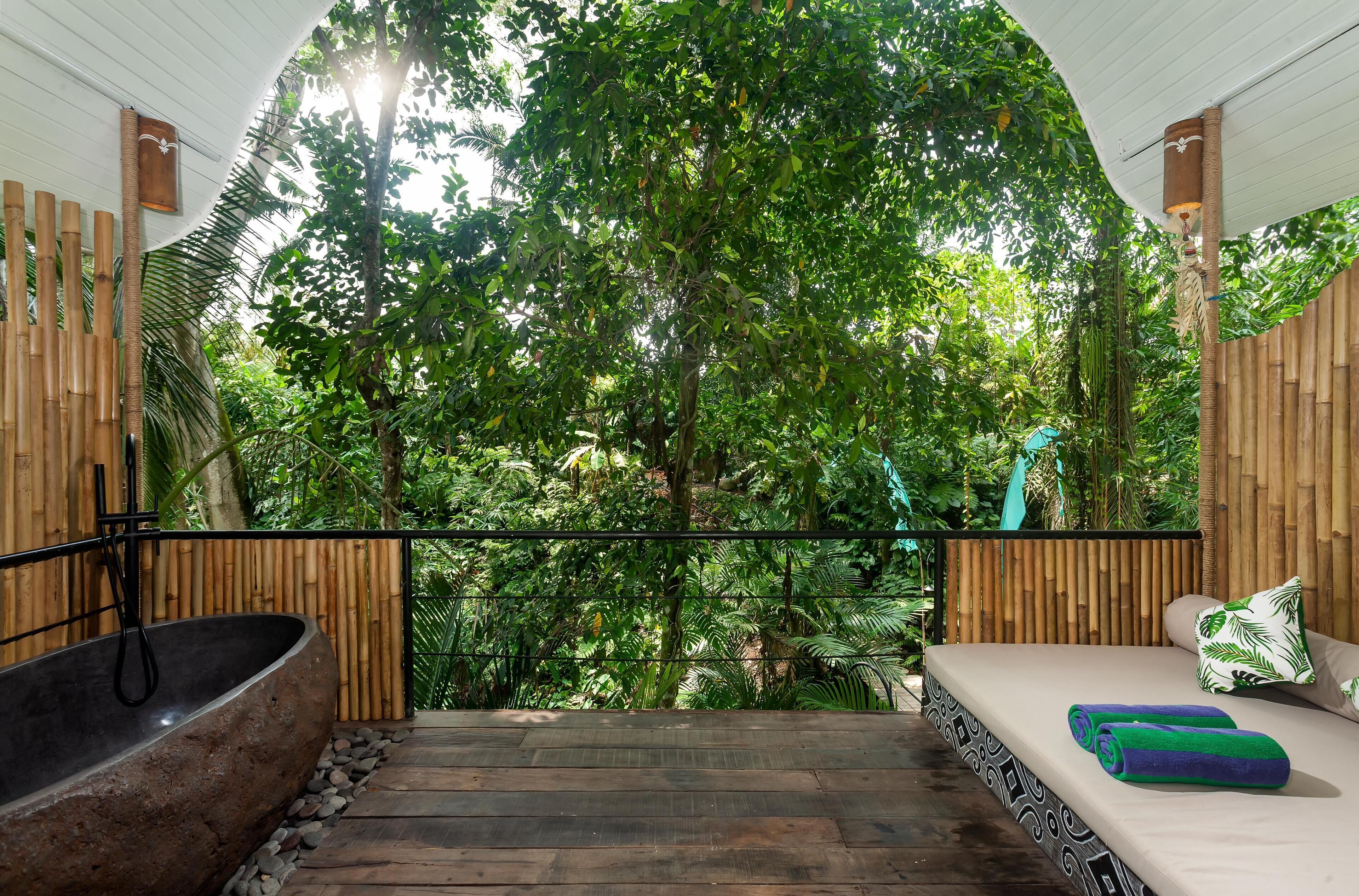 A wooden deck with a stone bathtub, a bed with green and white pillows, and a view of dense green foliage.