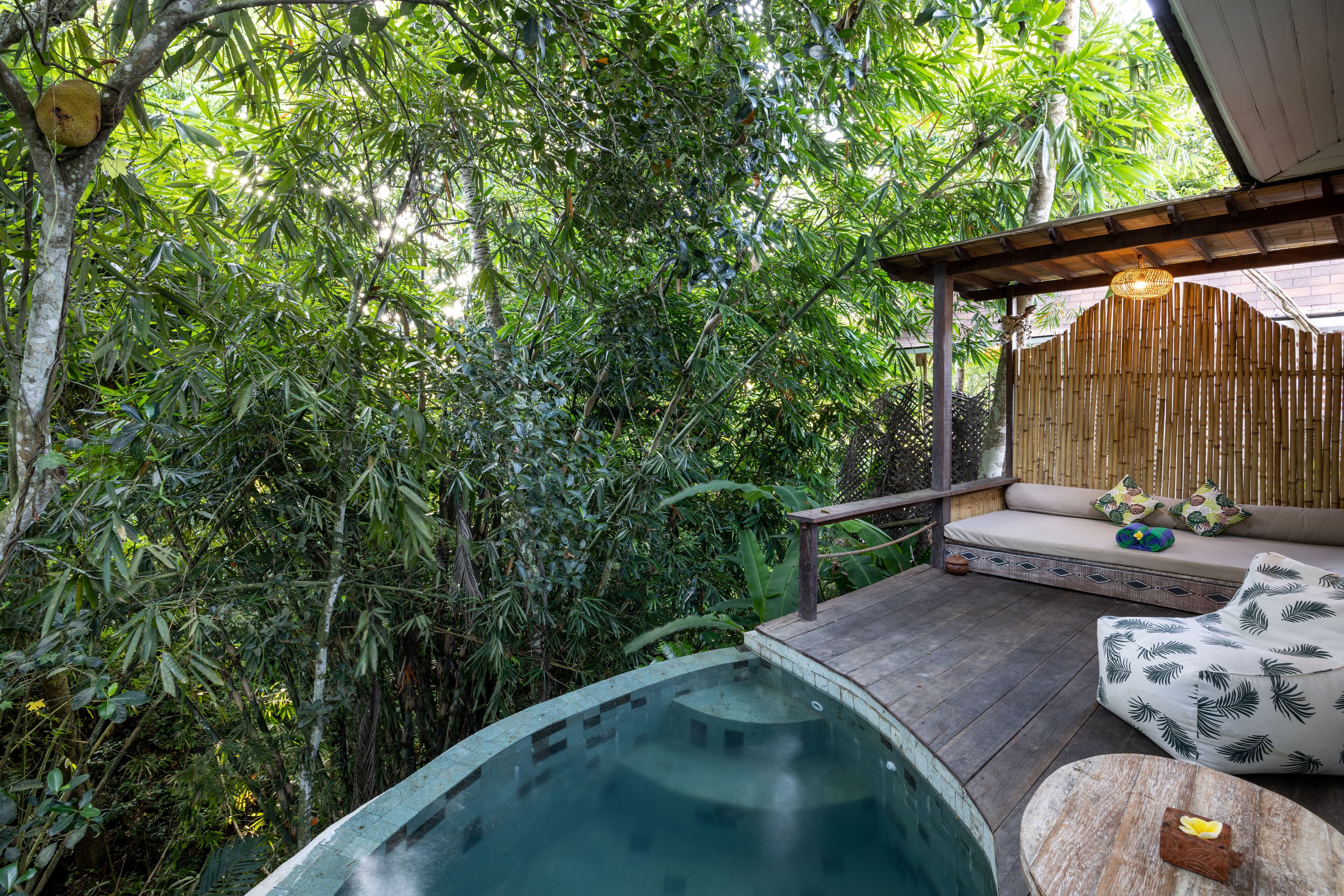 Un área de piscina con un deck de madera, una sala de estar con banca acolchada y abundante vegetación alrededor.
