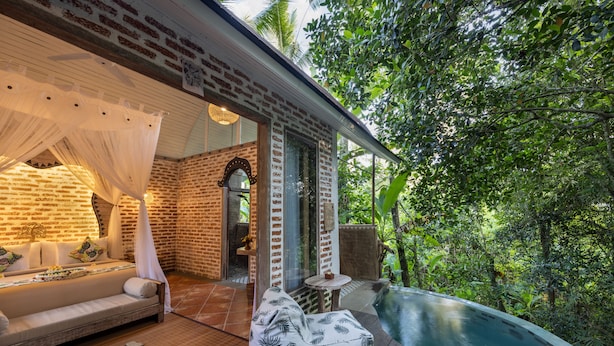 A cozy outdoor seating area with a sofa and cushions, a small table, and a bench, all set on a wooden deck with a view of a lush garden and a swimming pool. - Sacred Canyon Ubud by GenuineHost (Payangan)
