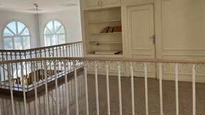Une pièce spacieuse avec un plafond haut, des murs blancs et un balcon doté d’une rambarde en métal. De grandes fenêtres laissent entrer la lumière naturelle, et un meuble blanc avec des étagères est adossé au mur.