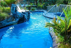 Piscina con caída de agua, rodeada de plantas tropicales y una terraza de madera.