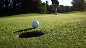 Une balle de golf sur un green, près d’un trou, et une personne à l’arrière-plan.