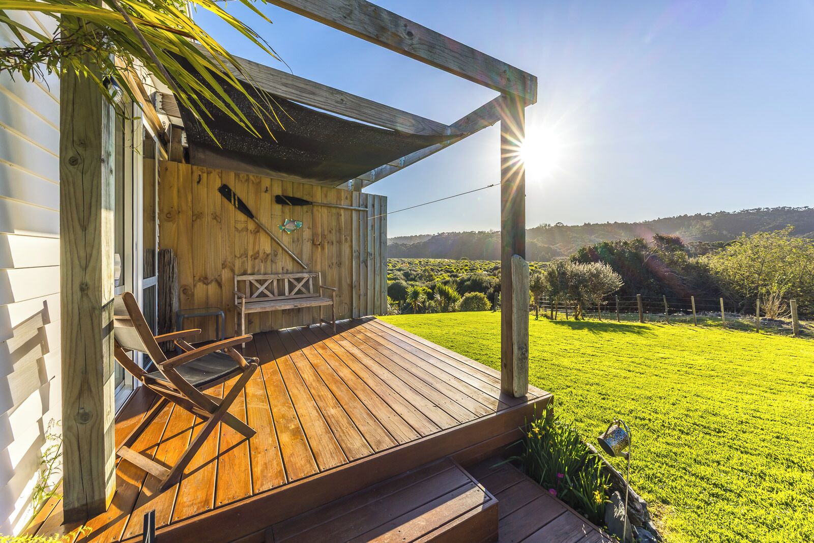 A wooden deck with a bench and chairs, a pergola, and a view of a green field and hills.