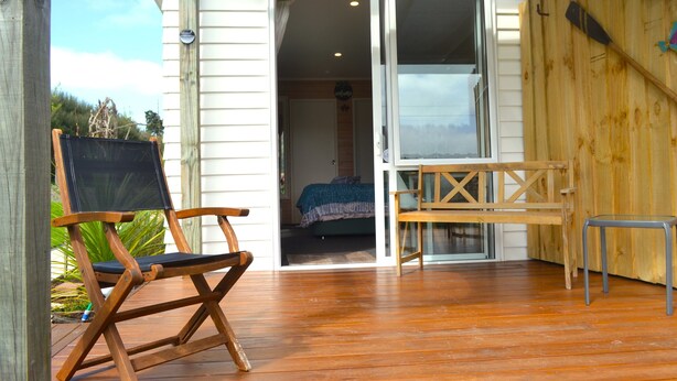 Une terrasse en bois avec une chaise pliante, une petite table et une porte-fenêtre coulissante donnant sur une chambre à coucher équipée d’un lit et d’une table de chevet.