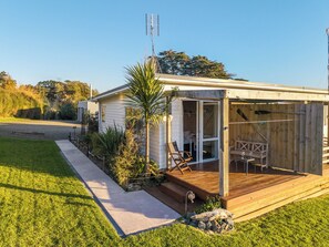 A single-story house with a covered porch, a sliding glass door leading to an outdoor seating area, and a small garden with a palm tree. - Manuka Views - Close to Thermal Hot Pools (Kaukapakapa)