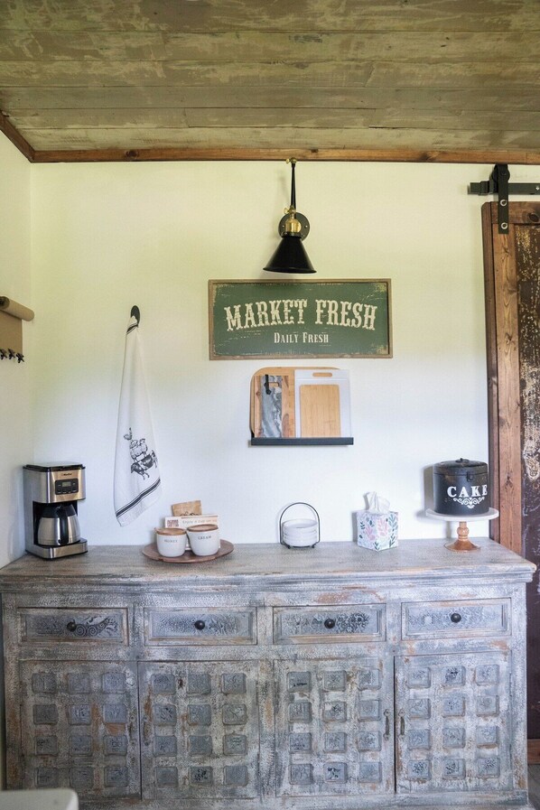 A rustic kitchen counter with a coffee maker, a hanging cloth, a vintage sign, and a cake display.