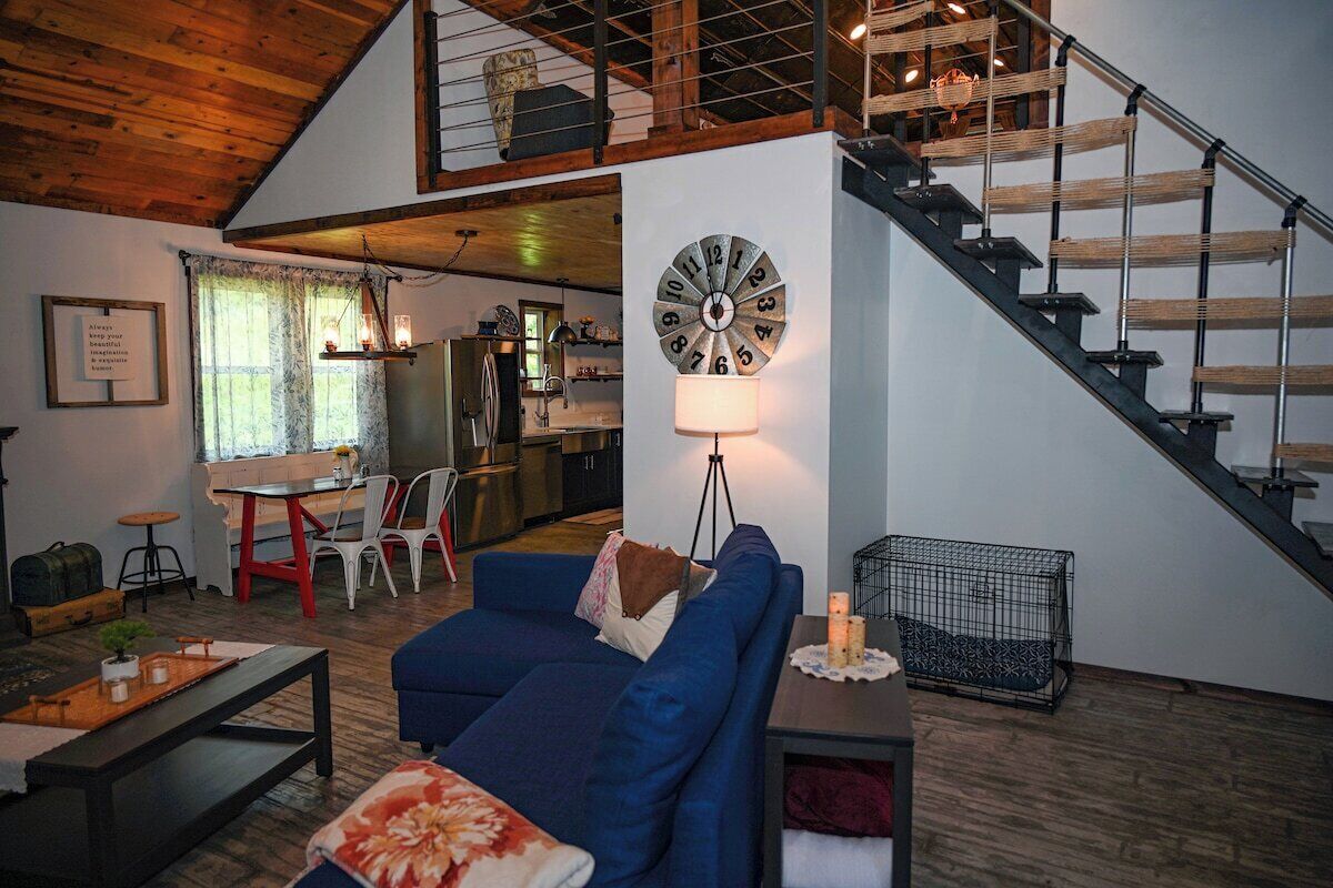 A modern living room with a blue sofa, a wooden coffee table, and a dining area with a red table and chairs. A staircase with a metal railing leads to an upper level.