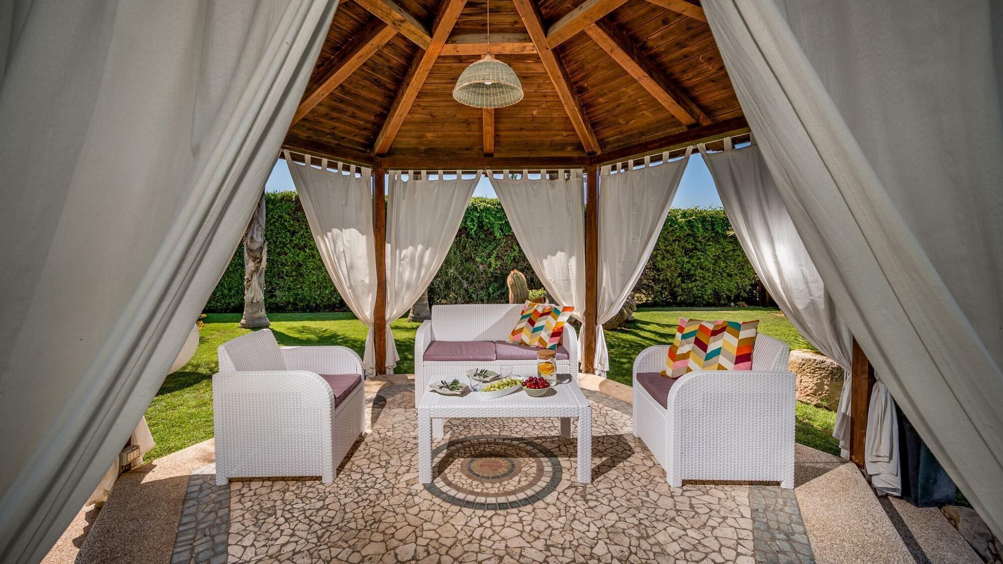 A covered outdoor seating area with wicker furniture and a table set for dining, surrounded by white curtains and a wooden ceiling structure.