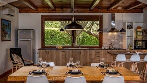 A modern kitchen with wooden cabinets, a large window with a view of greenery, and a dining area set for six with white chairs. - Villa Cutrera 6 Emma Villas (Chiaramonte Gulfi)