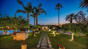 A well-maintained garden with a stone pathway leading to a building, surrounded by palm trees and a pool. - Villa Cutrera 6 Emma Villas (Chiaramonte Gulfi)